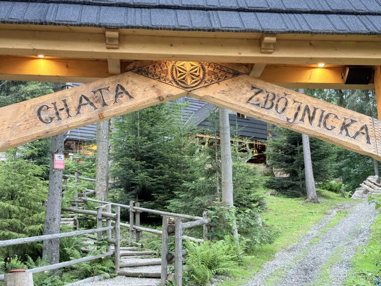 Drewniana brama wjazdowa z wyrzeźbionym napisem "Chata Zbójnicka", prowadząca żwirową ścieżką przez las w kierunku restauracji w Zakopanem.